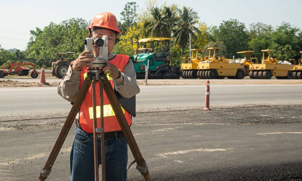 Land surveying work being done by a surveyor using a level instrument on a busy road construction project